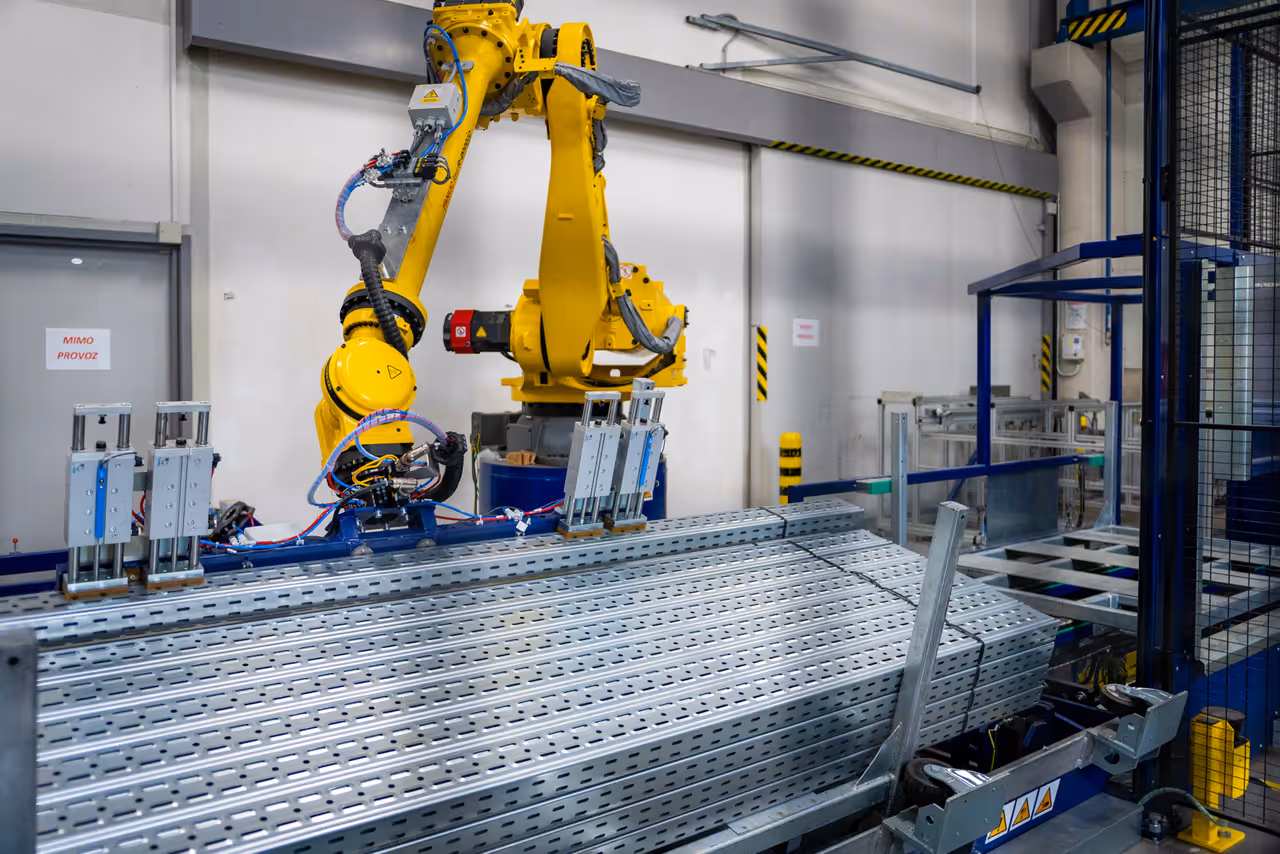 Žlutý průmyslový robot s manipulačním chapadlem nad stohem pozinkovaných perforovaných profilů